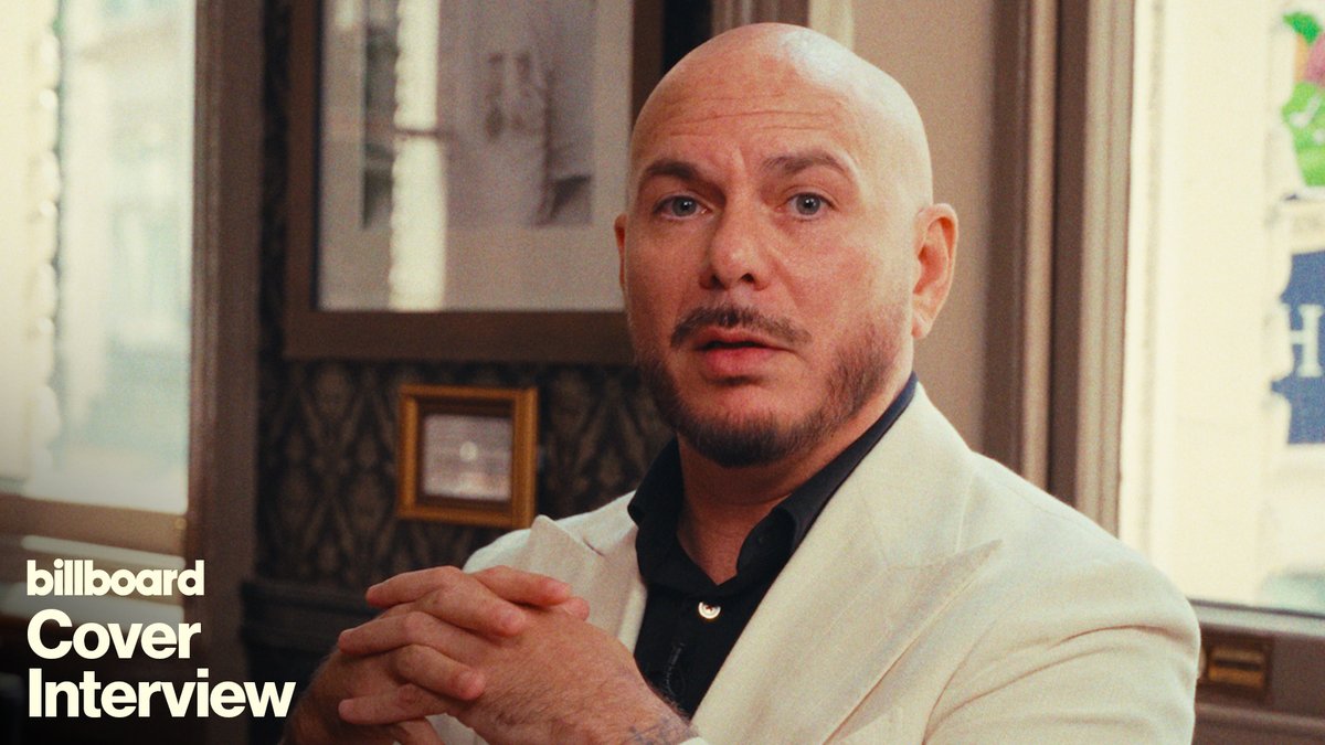 Billboard Cover Interview promotional image featuring Pitbull in a white suit jacket and black shirt, seated indoors with his hands clasped. The text “billboard Cover Interview” appears in the lower-left corner, accompanying a quote about how the Miami rapper’s multicultural upbringing shaped his approach to music ahead of his Billboard Latin Week Superstar Q&A.