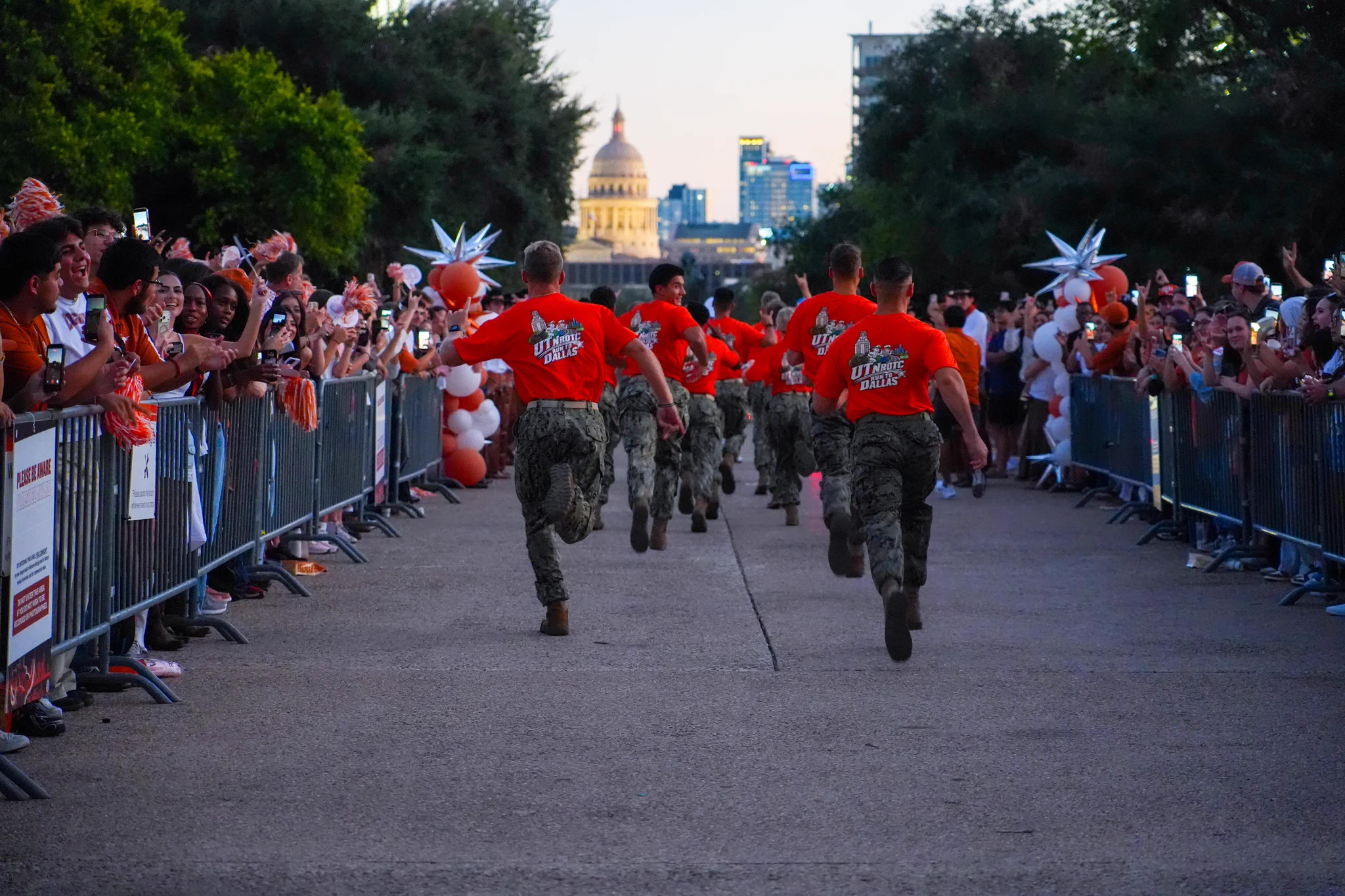 A group of men in red \"UTNROTC RUN TO DALLAS\" shirts and camouflage pants run away from the camera down a paved path. A diverse crowd cheers from behind metal barriers, many holding up phones. A city skyline with a prominent domed building is visible in the distance at dusk.