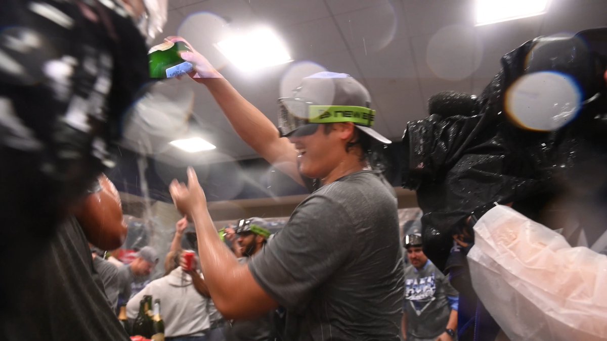 A baseball player wearing a "REMOVE" green headband and backwards cap celebrates in a locker room, raising a green beer can amid a shower of spraying liquid while teammates cheer around him, capturing the jubilant postseason celebration referenced in the caption about Shohei Ohtani enjoying #Postseason moments.