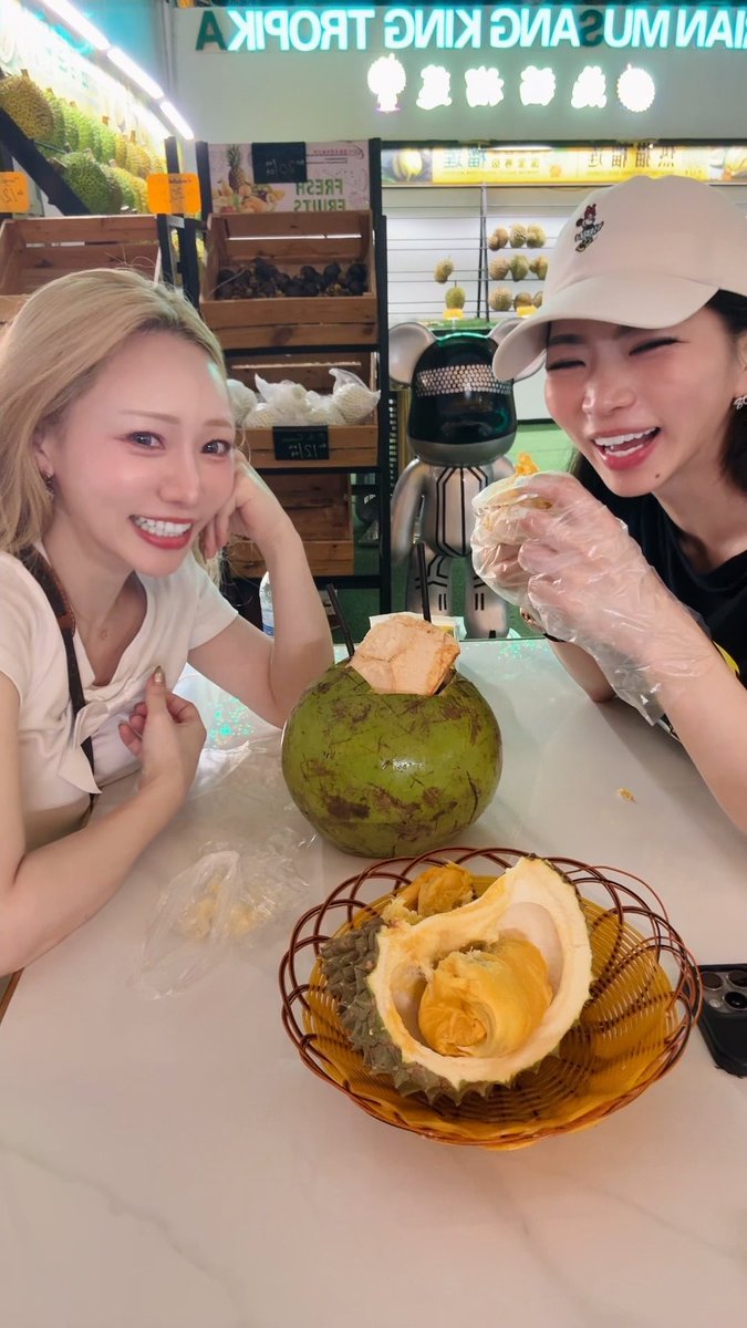 Two women share a joyful first encounter with a fresh durian at "Love King Troika" market, with one wearing a plastic glove and holding a piece of the spiky fruit while the other poses beside it. A basket of the opened durian sits on the counter between them, capturing the playful "first durian" moment referenced in the post about Asamiyako trying the famously pungent fruit.