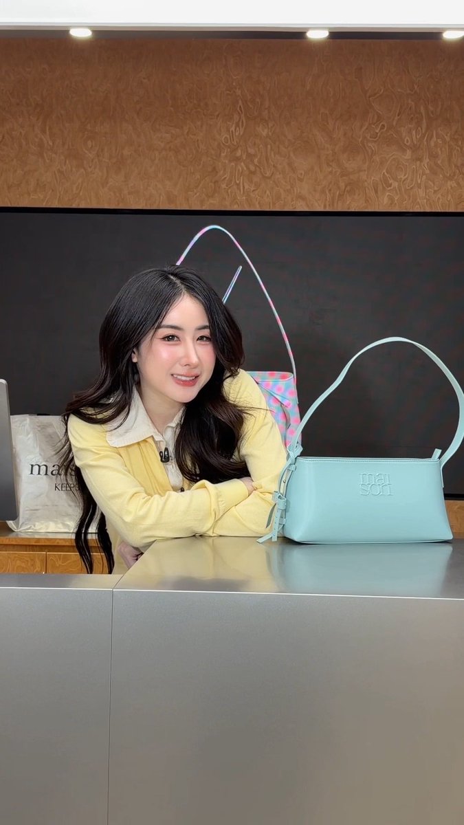 A smiling woman in a yellow cardigan leans on a counter next to a light blue handbag with 'manson' written on it, with a pink leopard-print bag visible behind her.
