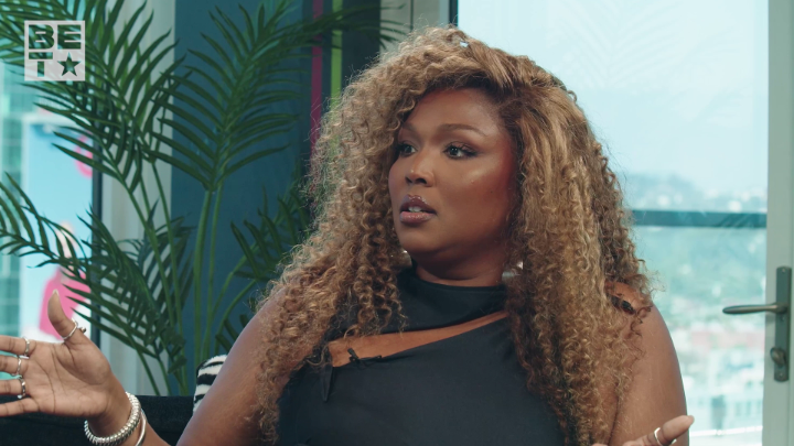 A screenshot from a BET network show featuring a woman with long, curly blonde hair wearing a black top, gesturing with her hands during an interview setting with a plant in the background.