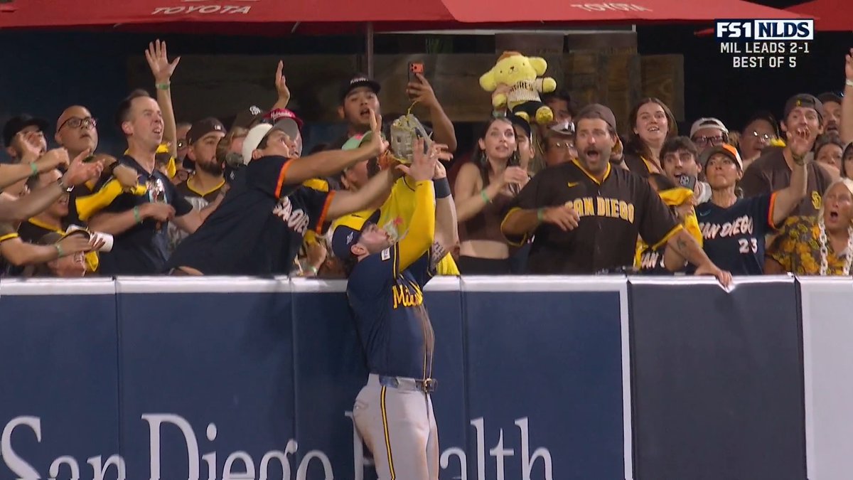 A San Diego Padres player leans over the outfield wall at San Diego Health Park, reaching up with his glove as fans in Padres gear react with outstretched arms, while the FS1 broadcast graphic shows "MIL LEADS 2-1 BEST OF 5" during the NLDS—matching the caption's reference to a near game-tying homer that fell just short of the fans' grasp.