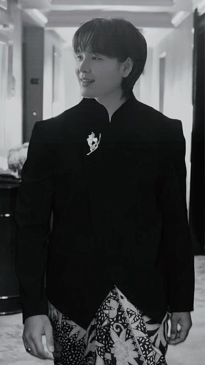 A black-and-white photo of a smiling East Asian man with short dark hair, wearing a black high-collared jacket with a decorative brooch and patterned pants.