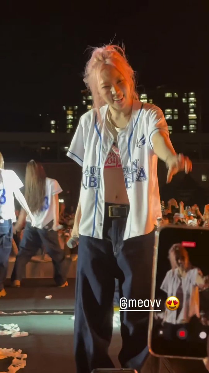 A young woman with pink hair wearing an open blue and white baseball jersey and baggy jeans, smiling and pointing downward on a stage during a concert, with city buildings visible in the background.