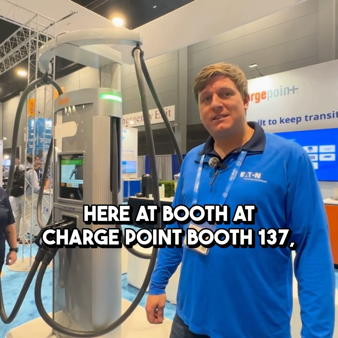 A man in a blue Eaton shirt stands beside an EV charger at a trade show with a caption reading 'HERE AT BOOTH AT CHARGE POINT BOOTH 137'.
