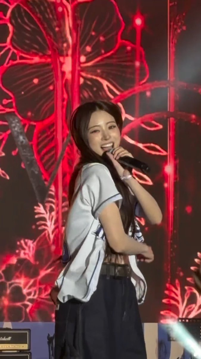 A young woman with long dark hair smiles while singing into a microphone on stage in front of a red LED backdrop.