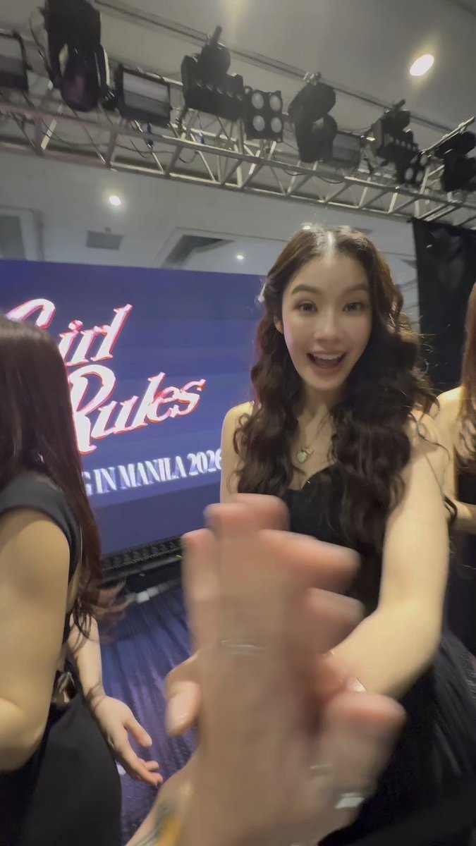 A young woman with long dark wavy hair and a black top smiles excitedly at the camera in what appears to be a backstage area, with a partially visible sign reading 'Rules' and 'IN MANILA 2026' behind her.