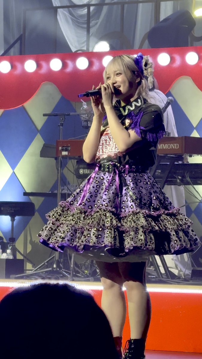 A young blonde performer singing into a microphone on stage, wearing an elaborate purple and black ruffled dress with a leopard print pattern, with a Yamaha keyboard visible behind her.