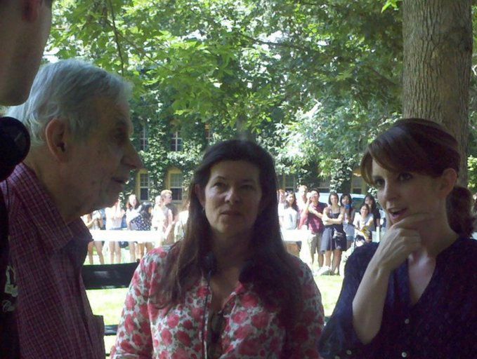 A candid outdoor photo shows an elderly man, a woman with dark hair in a floral shirt, and a woman with auburn hair engaged in conversation, with a crowd of people visible in the background.