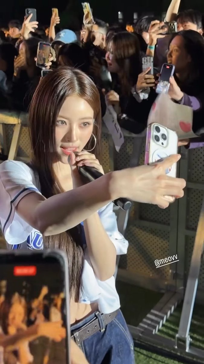 A woman with long brown hair holds a microphone and a phone, performing for a crowd of fans taking photos at a concert (watermark: @meovv).