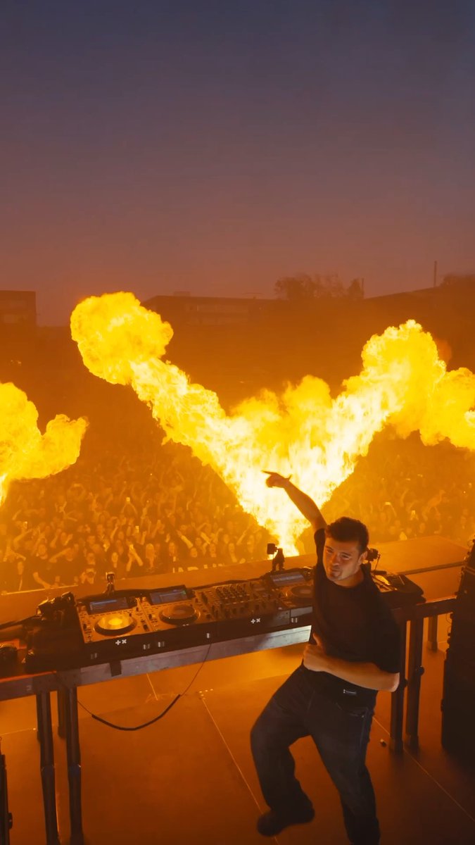A DJ performs on stage in front of a massive crowd, with dramatic flames erupting in a V-shape behind him during a concert at dusk.