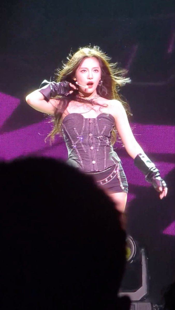 A concert photo of a female performer with long wavy hair, wearing a dark corset-style outfit and long gloves, singing into a headset mic against a purple-lit background.