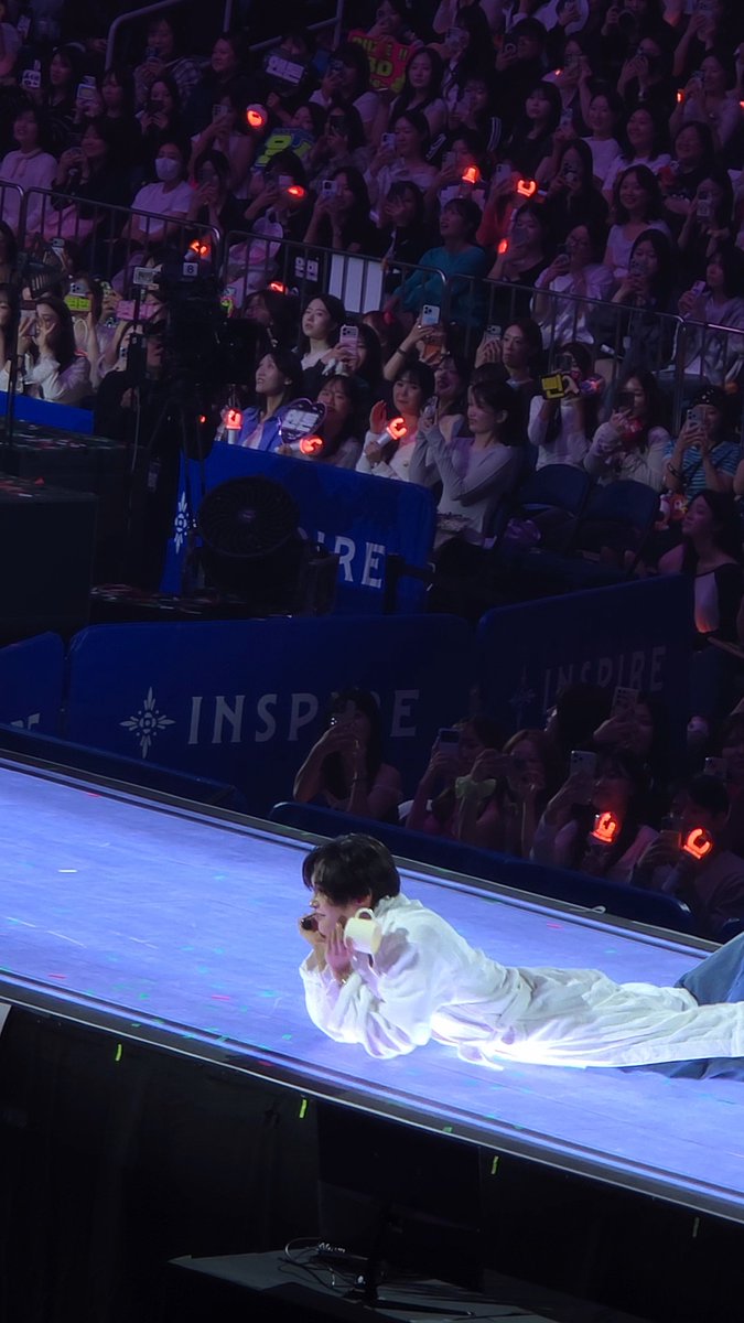 A performer in white clothing lies on a stage illuminated with blue light during a concert, while an enthusiastic audience behind a barrier holds up phones and red heart-shaped light sticks.
