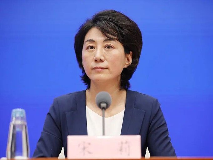 A woman in a dark blazer sits at a podium with a microphone and a nameplate reading '宋莉' (Song Li) against a solid blue backdrop.