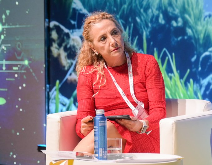 A woman with curly blonde hair wearing a red ribbed dress sits in a white armchair on a stage, holding a phone and a small notebook; a water bottle and glass are on the table in front of her.