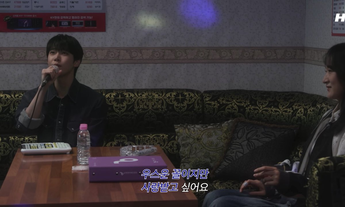 A scene from a Korean karaoke room showing a man in a dark shirt holding a microphone and singing, with another person seated beside him, featuring Korean subtitles "우선은 끝이지만 사랑받고 싶어요" (It ends first, but I want to be loved).