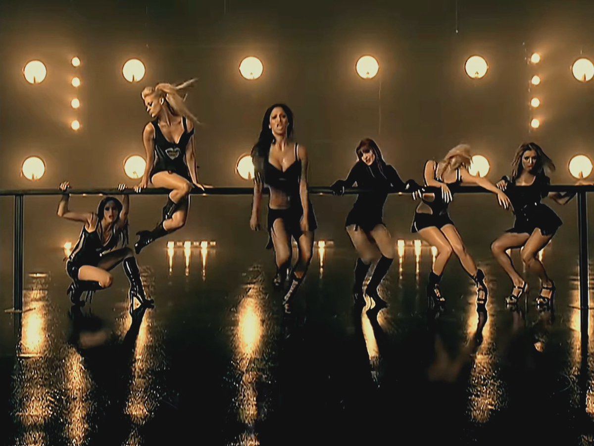 Seven women in black outfits perform a choreographed dance on a stage with a bar-like railing, lit by warm circular lights in the background and a reflective floor.