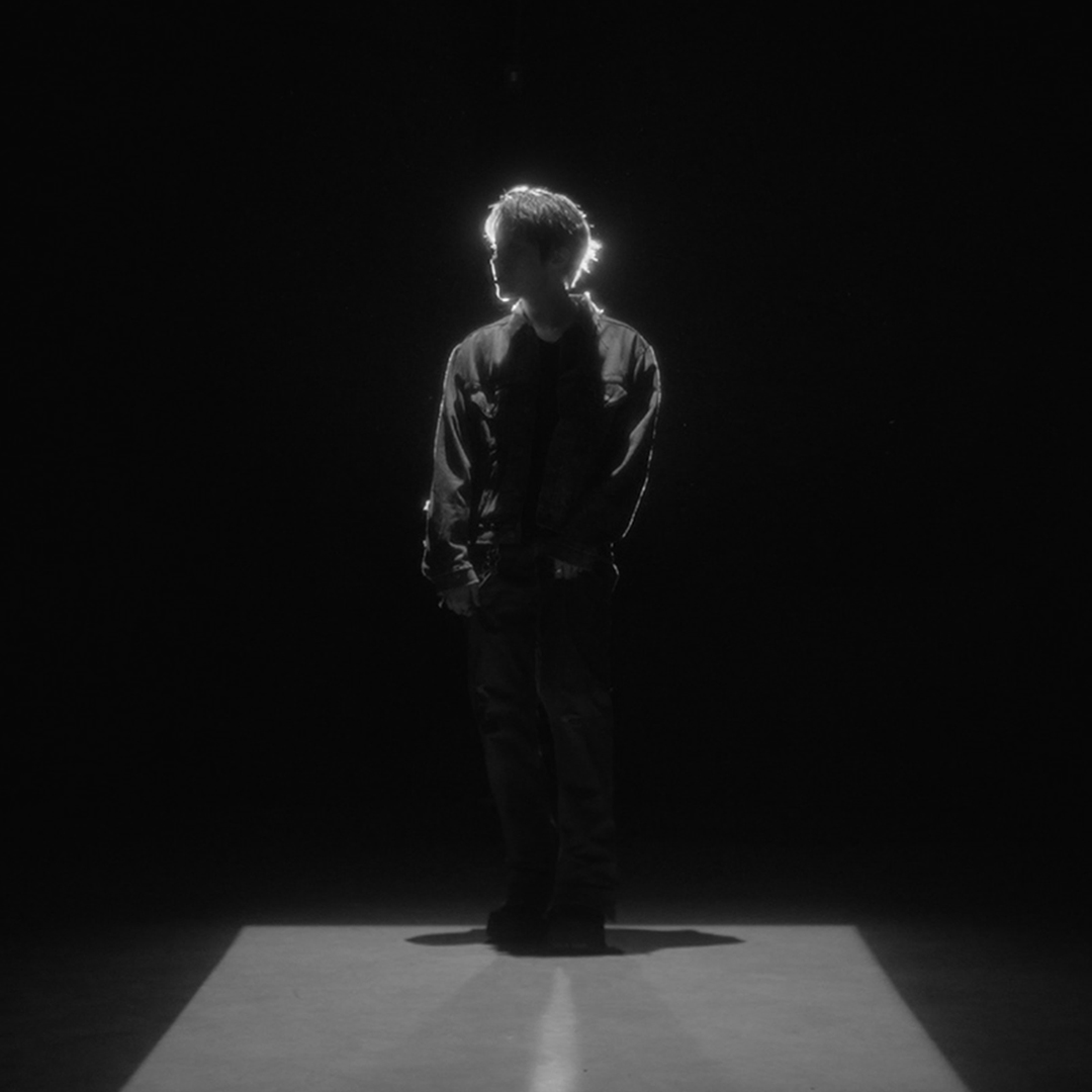 A dramatic black and white photograph of a young man standing in a spotlight with his back to the camera, hands in his pockets, surrounded by complete darkness.