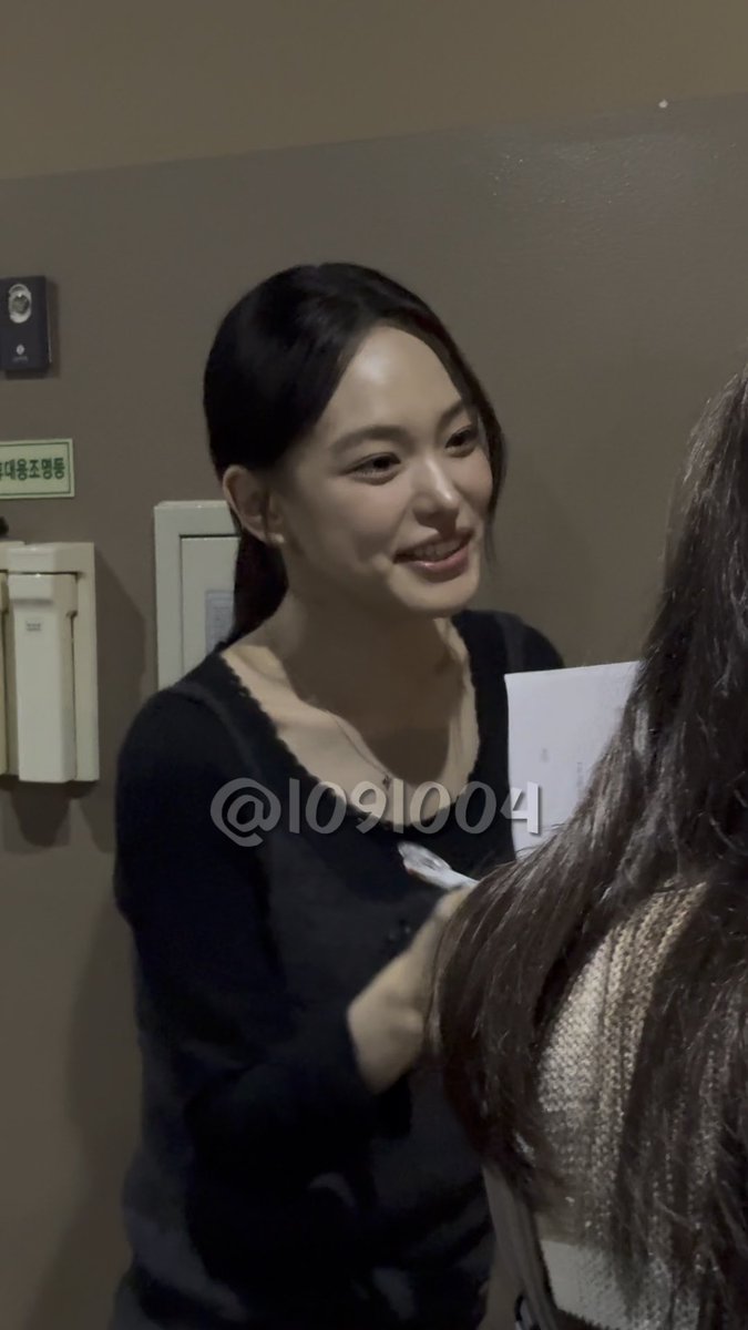 A photo of a smiling East Asian woman with dark hair pulled back, wearing a black top, appearing to sign autographs or interact with someone, watermarked '@1091004'.