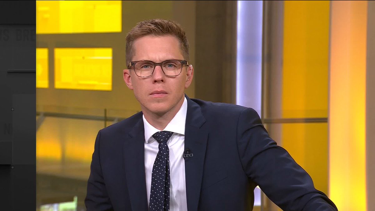 A male news anchor with blonde hair and glasses, wearing a dark suit and polka-dot tie, sits at a desk in a yellow-lit broadcast studio, appearing to deliver the breaking report about Russian authorities denying reports of a second plague case in their country.