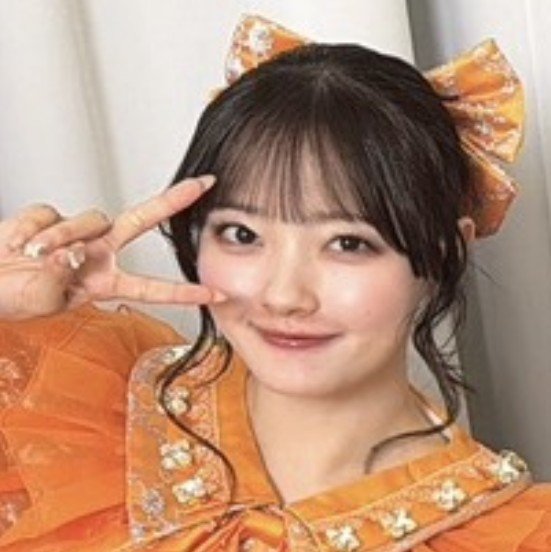 A young woman wearing an orange outfit with floral details and an orange hair bow makes a peace sign near her face.