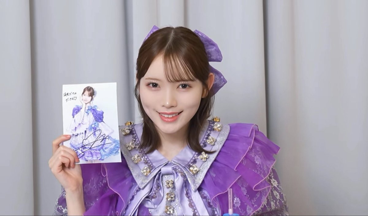A smiling young woman with brown hair wears a detailed purple outfit with ruffled shoulders and a large hair bow. She holds up a signed photograph of herself in a similar purple dress, with handwritten text on the photo.