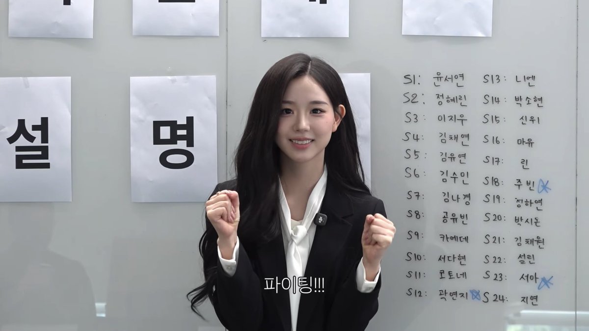A young woman with long dark hair smiles and raises her fists in front of a board displaying Korean text, including names like "윤서연," "정혜리," and "이시우" numbered S1 through S24, with some names crossed out.
