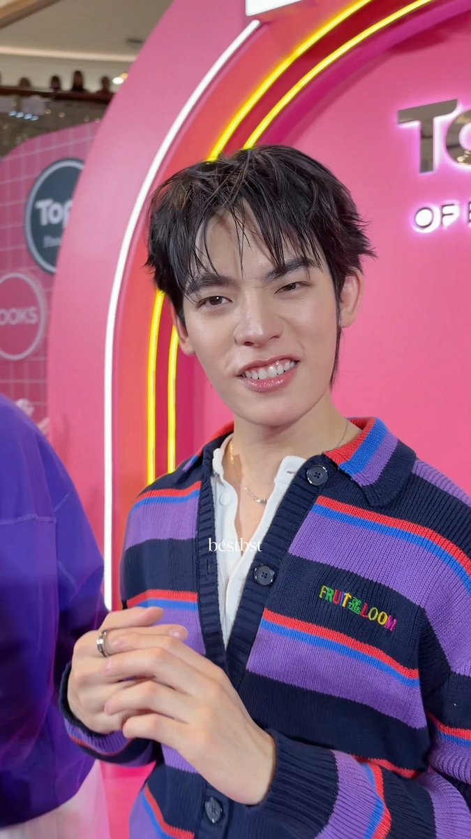 A young man with dark hair smiling, wearing a colorful striped "FRUIT OF THE LOOM" cardigan over a white shirt, posing in front of a bright pink neon sign at a store event.