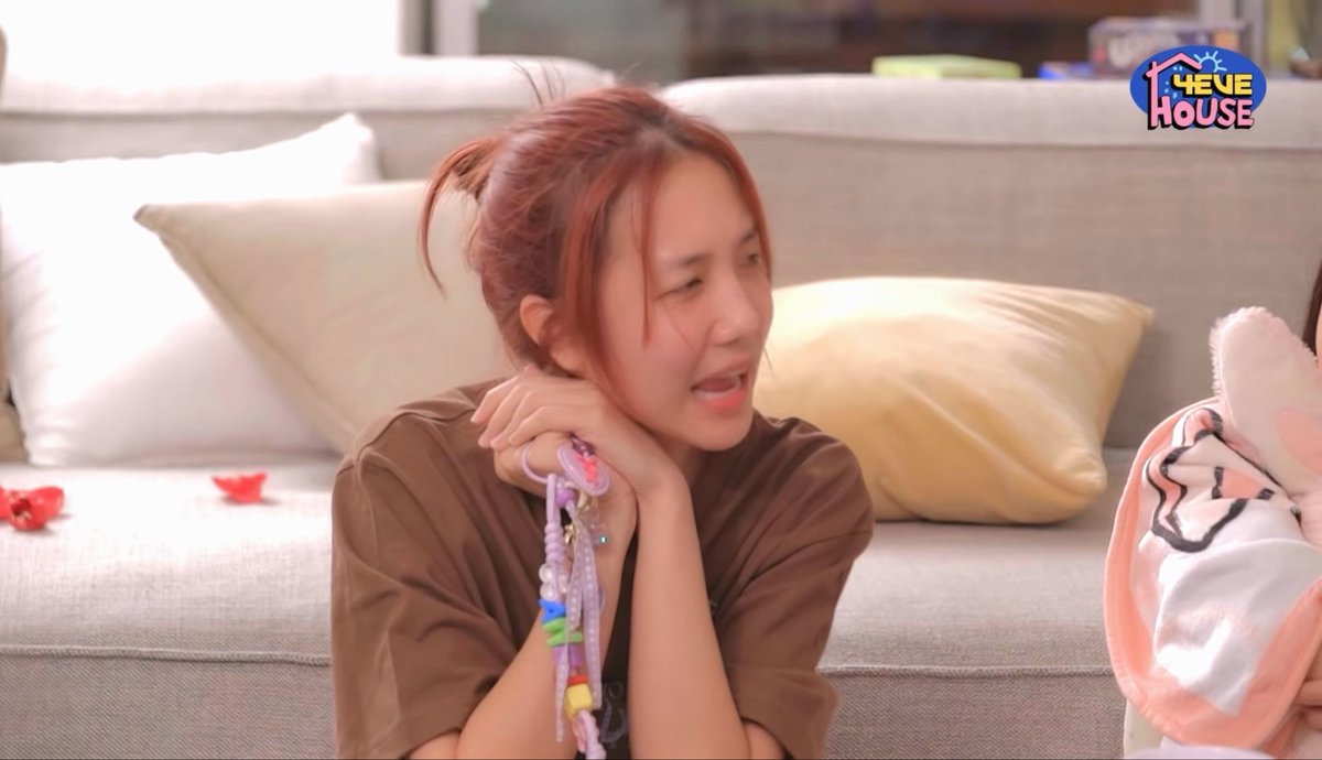 A woman with reddish-brown hair in a brown top sits on a beige couch, holding a colorful keychain while chatting with another person holding a pink cap. The "Thevie House" logo appears in the top right corner, matching the playful Thai slang caption about following Jo's lead.