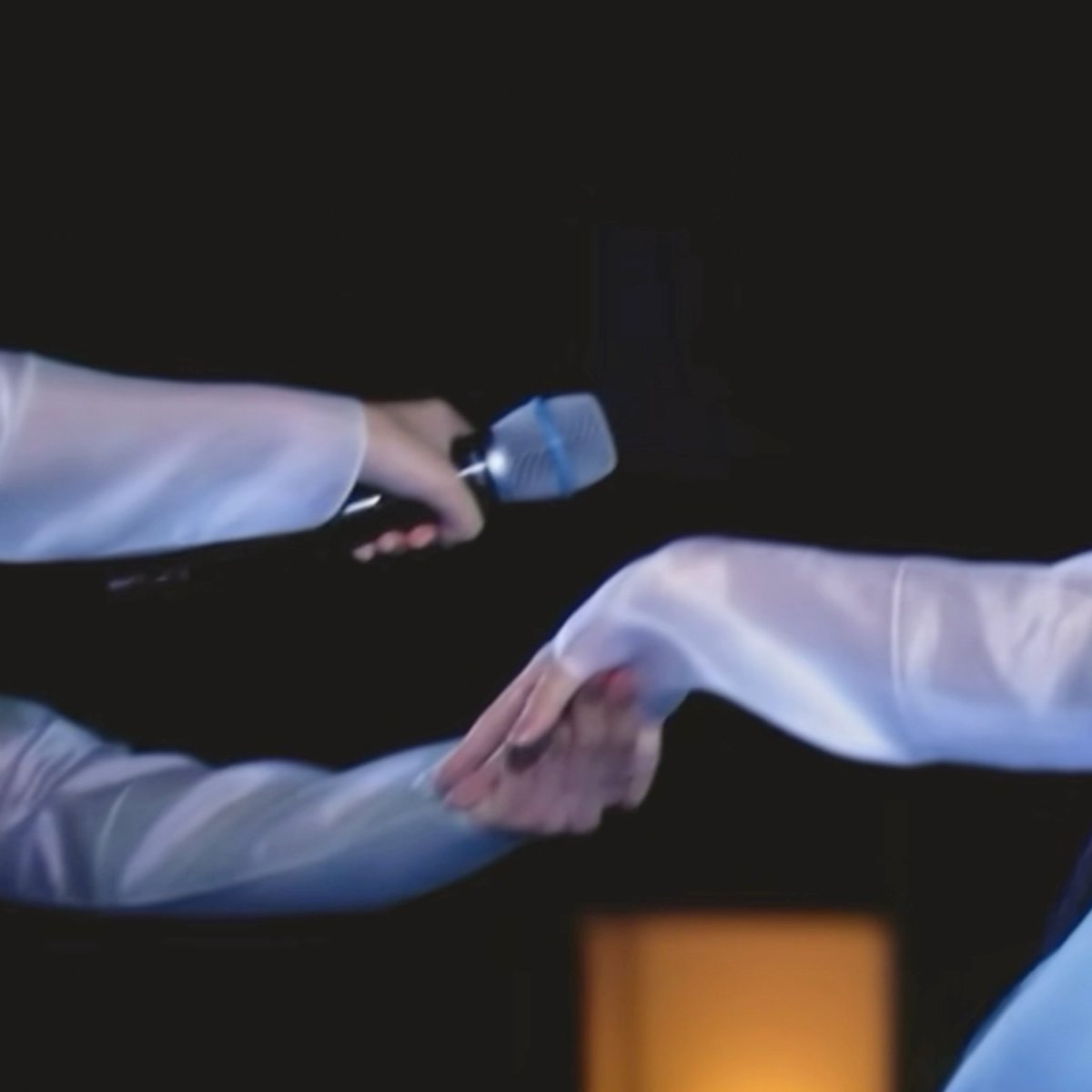 Two people wearing white garments appear to be exchanging or touching hands in a dark setting, with one person holding a small flashlight.