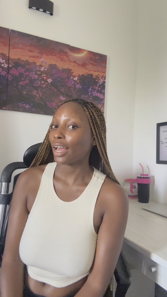 A young woman with long braided hair wearing a cream-colored tank top sits in an office chair, with a cherry blossom sunset painting on the wall behind her.