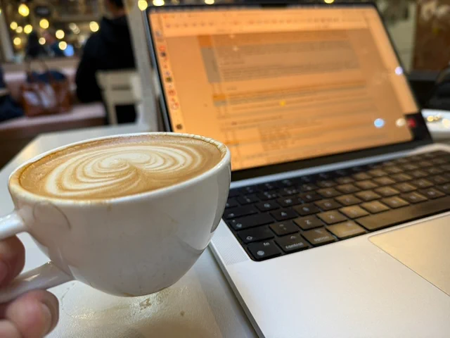 A hand holds a white cup of coffee beside an open laptop on a café table. The laptop screen and warm background lights are out of focus.