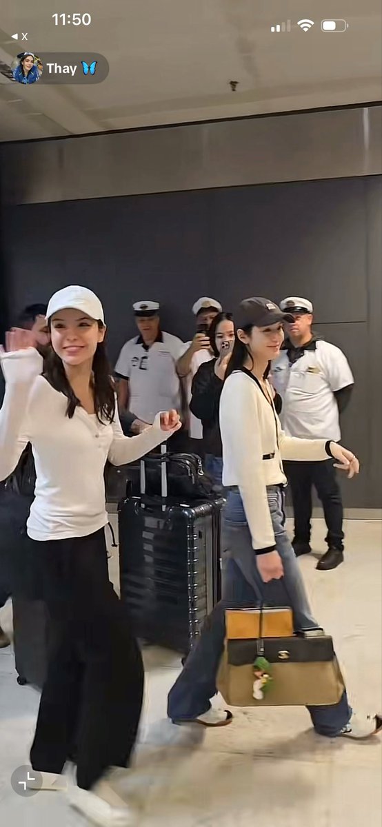 A screenshot from a video shows two smiling women in white tops walking through an airport or similar location with luggage, accompanied by uniformed security personnel, with a contact labeled 'Thay 🦋' at the top.