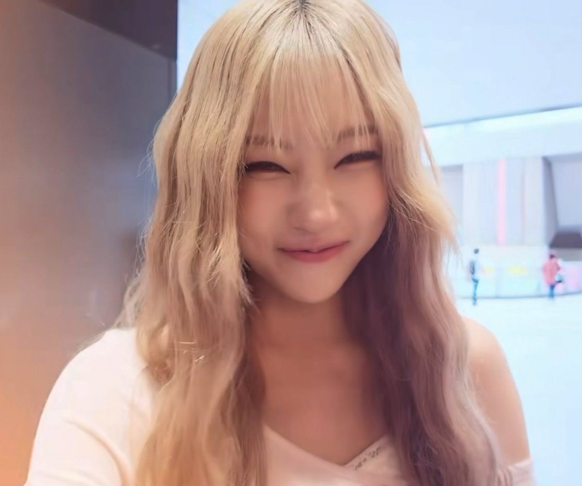 A close-up selfie of a smiling young woman with long blonde hair and bangs, standing in what appears to be an indoor public space or mall.