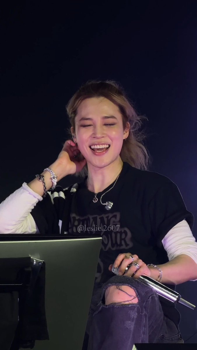 A smiling young woman with a ponytail wearing a black graphic t-shirt, ripped jeans, and multiple rings and bracelets, sitting at what appears to be a concert or event, holding a microphone.