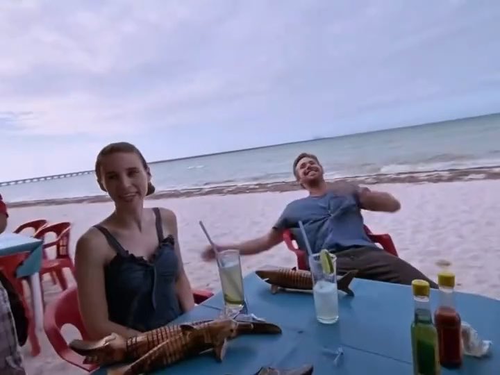 Two people relaxing at a beachside table with drinks, where the man is laughing and leaning back while the woman smiles at the camera, with a fish-shaped serving dish on the table.