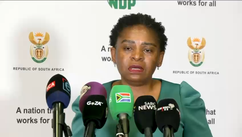 A South African government press briefing is shown, with a woman speaking at a podium backed by logos and text including “NDP,” “works for all,” “REPUBLIC OF SOUTH AFRICA,” and “A nation that works for all.” Several microphones are visible in front of her, including ones marked “GOZA TV,” “SABC NEWS,” and “AGT,” matching the accompanying post about an Inter-Ministerial Committee briefing on migration and enforcement of asylum laws.