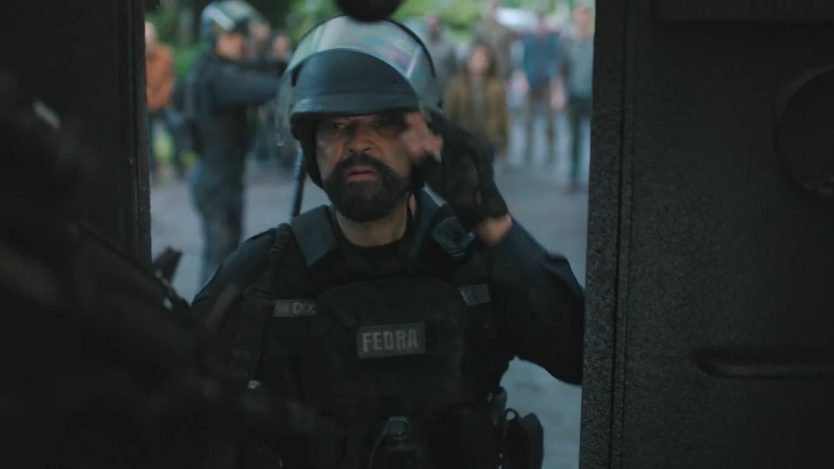A FEDRA soldier in riot gear and helmet stands at a doorway, adjusting his equipment, with a blurry crowd visible in the background. The "FEDRA" insignia on his chest armor suggests this is a scene from *The Last of Us*, where the militarized faction monitors civilians in a quarantine zone, likely prompting amusement due to the officer's serious demeanor amid a tense situation.