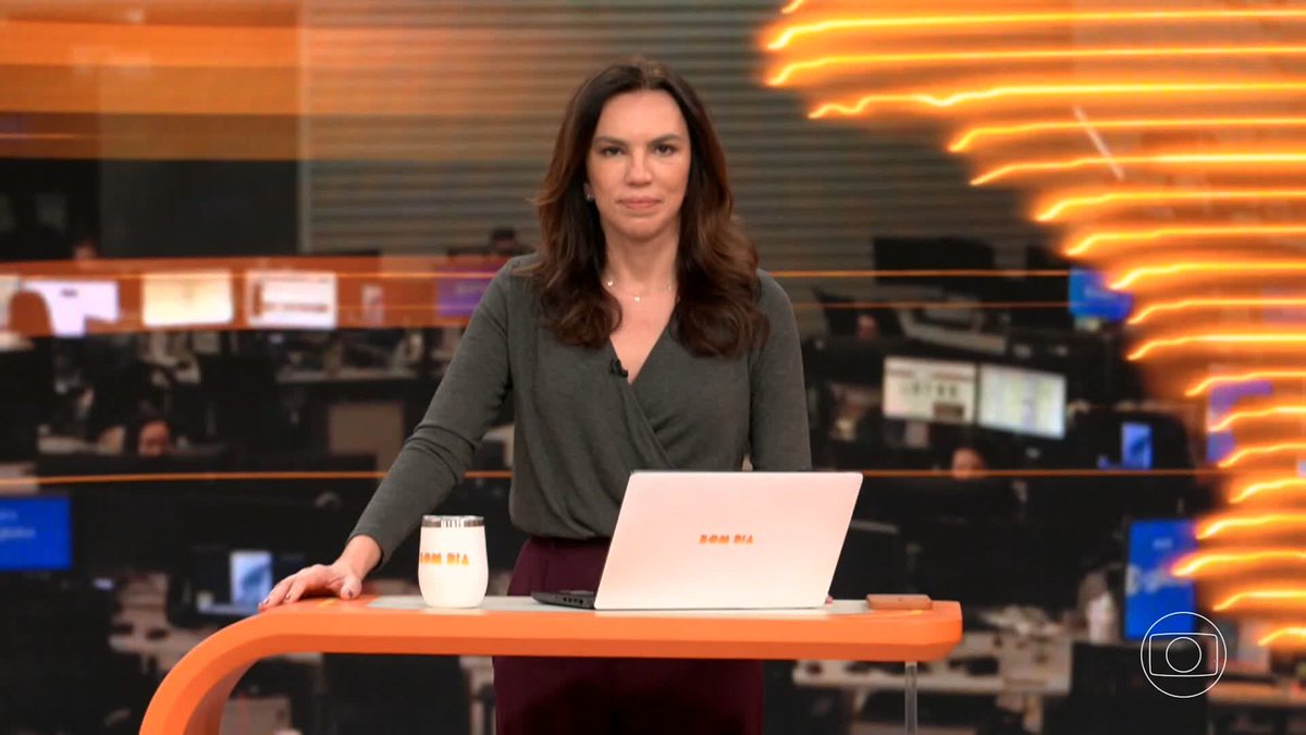 A female news anchor stands at a desk in a studio with an orange backdrop, behind a laptop labeled "BOM DIA" and a matching tumbler.
