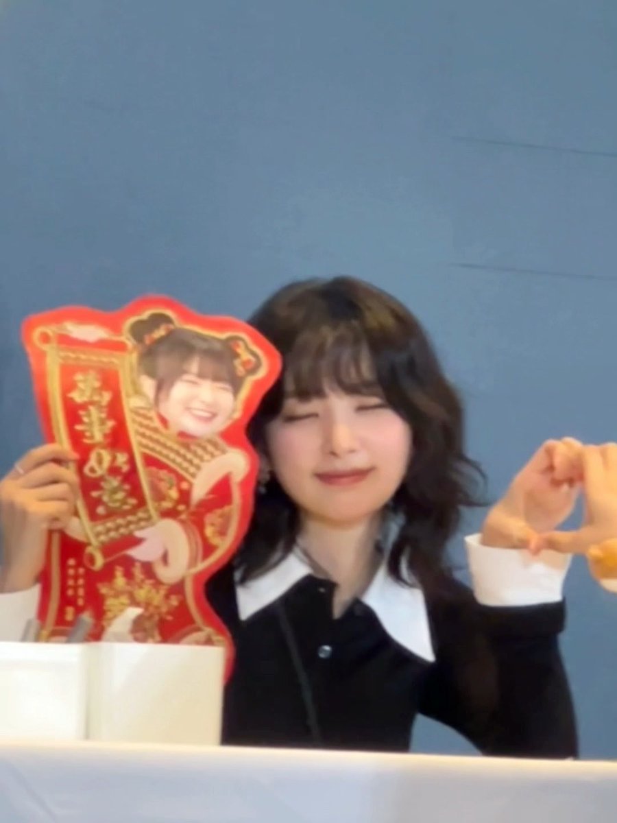 A smiling woman with dark hair in a black and white collared shirt holds a red Chinese decoration featuring a cartoon character and the text '万事如意'.