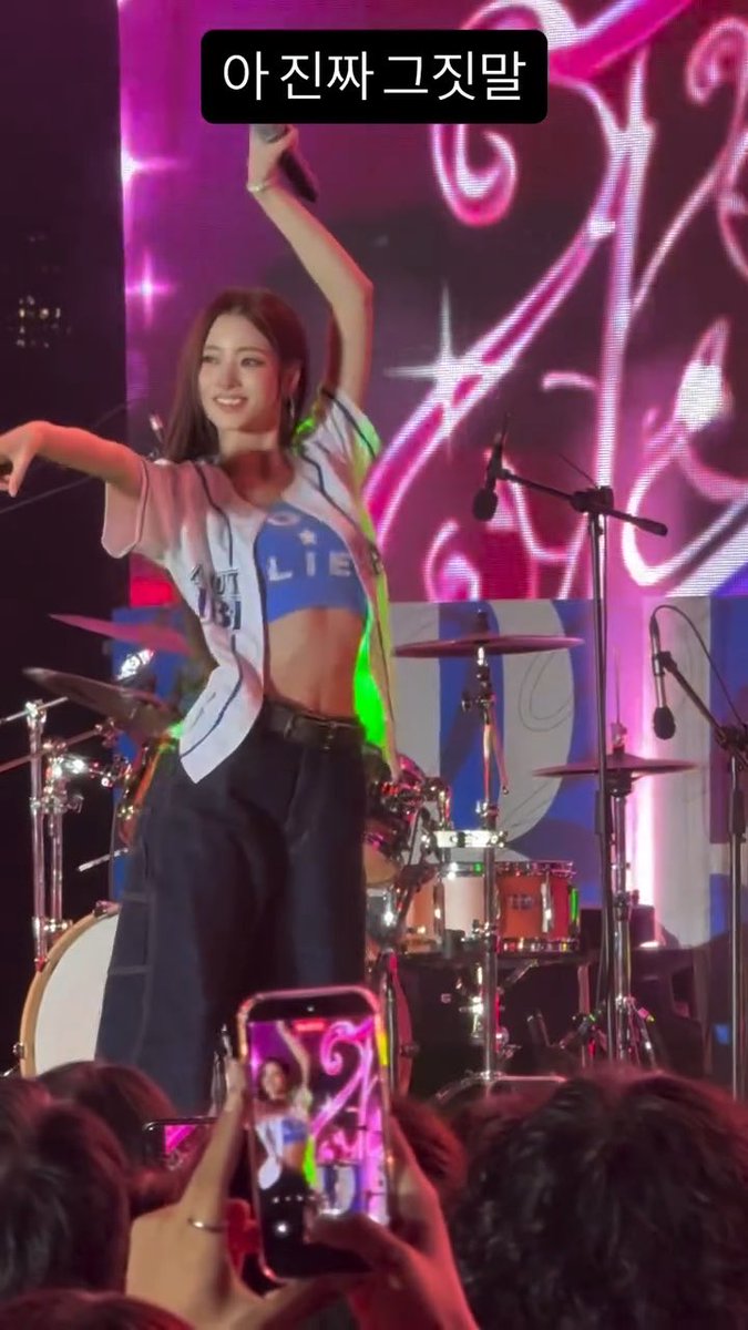 A K-pop idol performing on stage with a microphone raised, wearing a blue crop top and oversized shirt, with Korean text "아 진짜 그짓말" (translation: "Ah really, what a lie") overlaid.