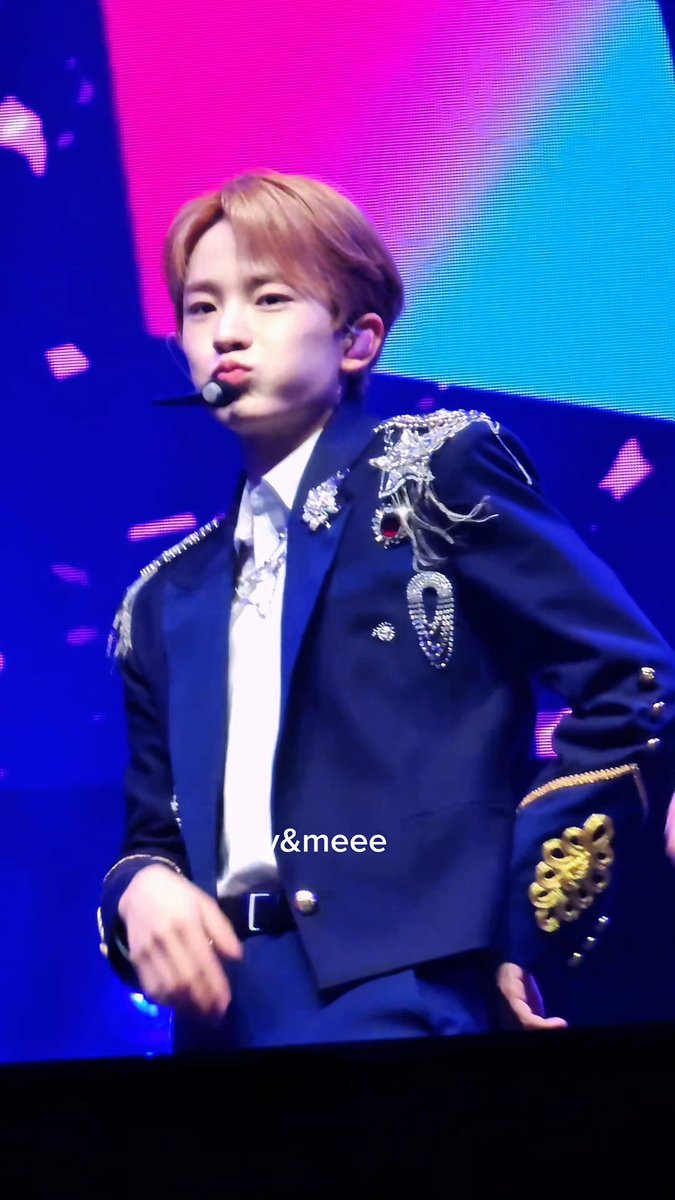 A performer with light brown hair wearing an ornate navy blue suit with silver embellishments and a headset microphone performs on stage with the watermark '&meee'.