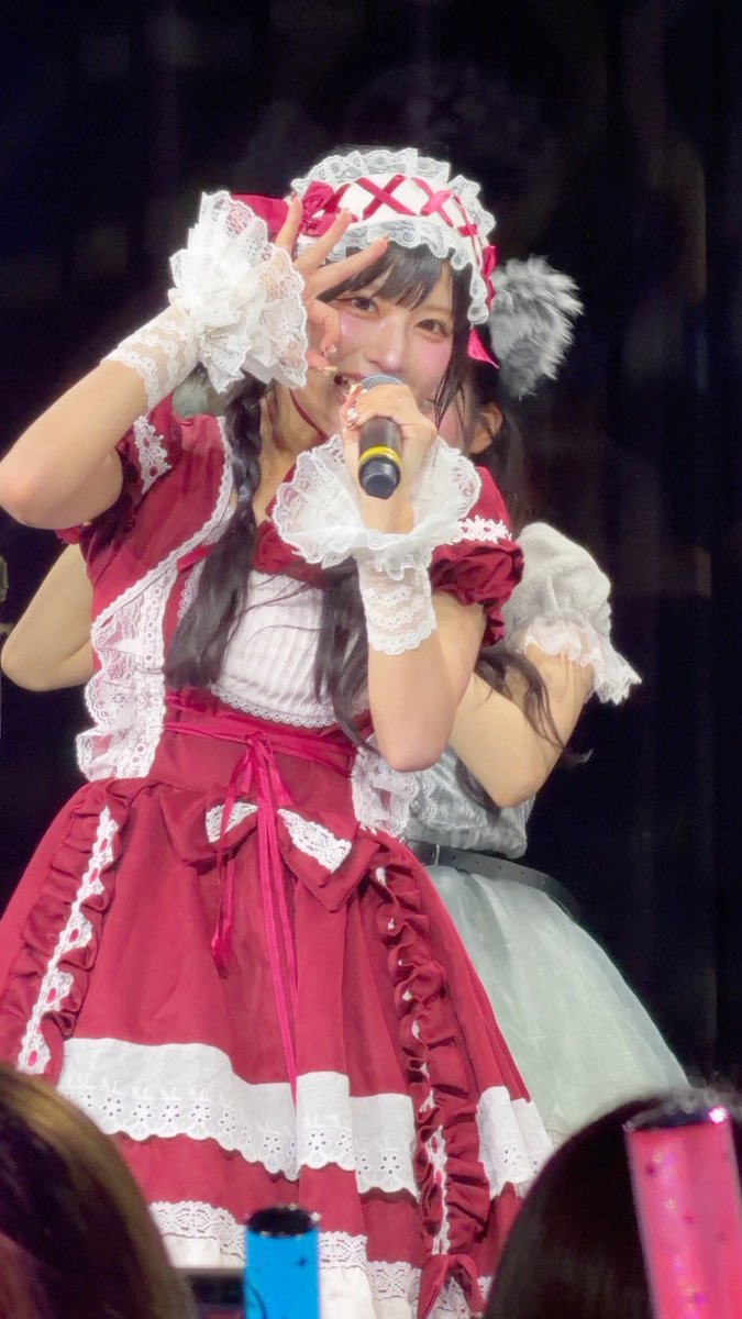 Two performers in elaborate red and white maid-style costumes perform on stage, with the front performer singing into a microphone and making a hand gesture.