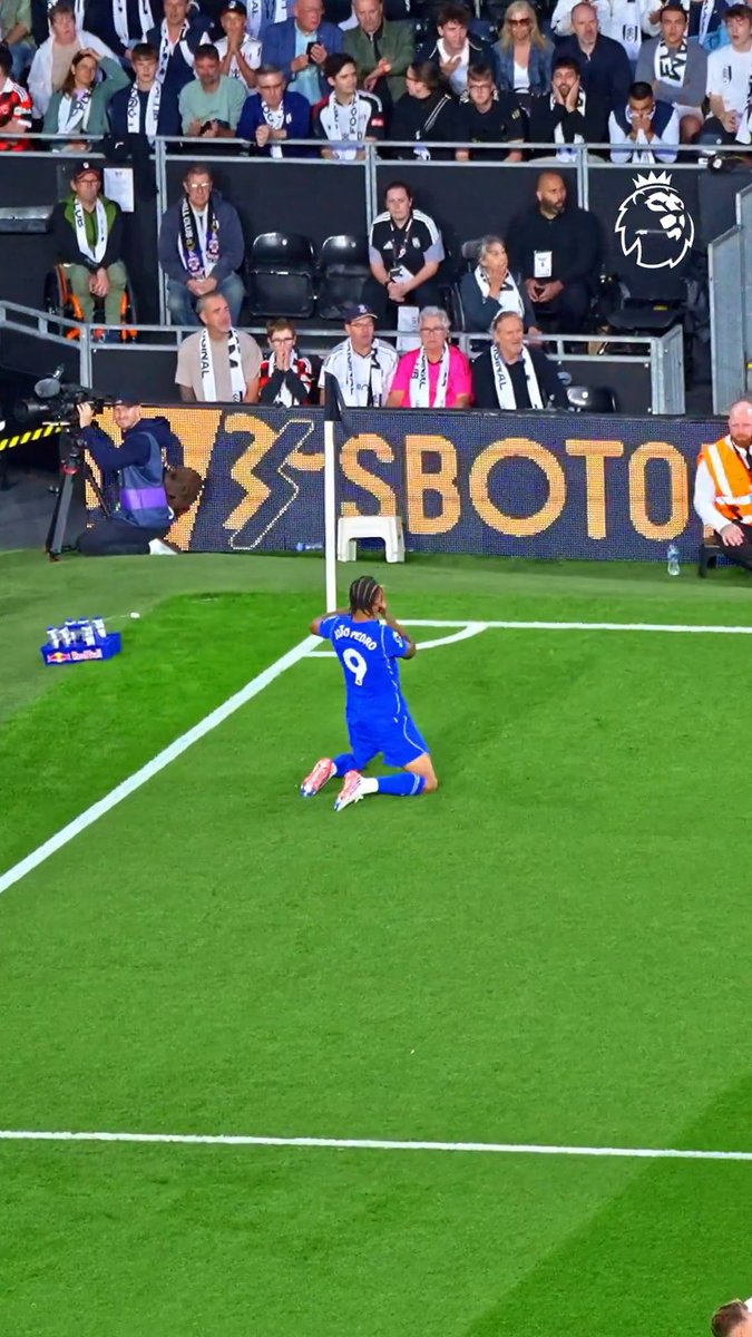 A Chelsea soccer player wearing jersey number 9 kneels on the grass near the corner flag, with 'SBOTOP' advertising visible on the boards and fans watching from the stands.