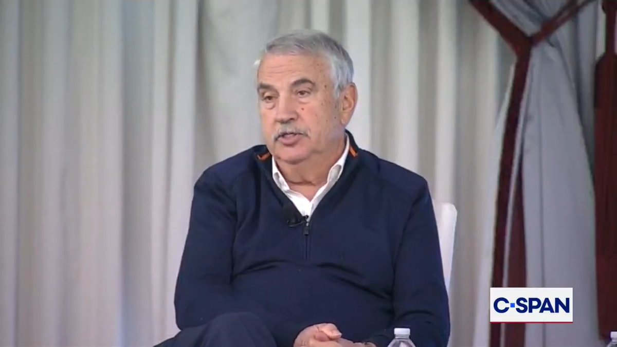 A gray-haired man with a mustache sits indoors in front of light curtains, wearing a navy sweater over a white collared shirt with a small microphone clipped near the zipper. The frame appears to be from a televised interview, with a white-and-blue “C-SPAN” logo in the lower-right corner and a plastic water bottle partly visible at the bottom.