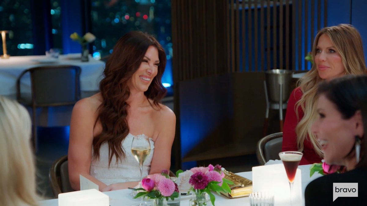 Women are seated at an elegantly set table with champagne, floral centerpieces, and a gold clutch, sharing a laugh during a glamorous dinner gathering. The Bravo logo is displayed in the bottom right corner, tying the scene to the Real Housewives of Orange County Tokyo episode.