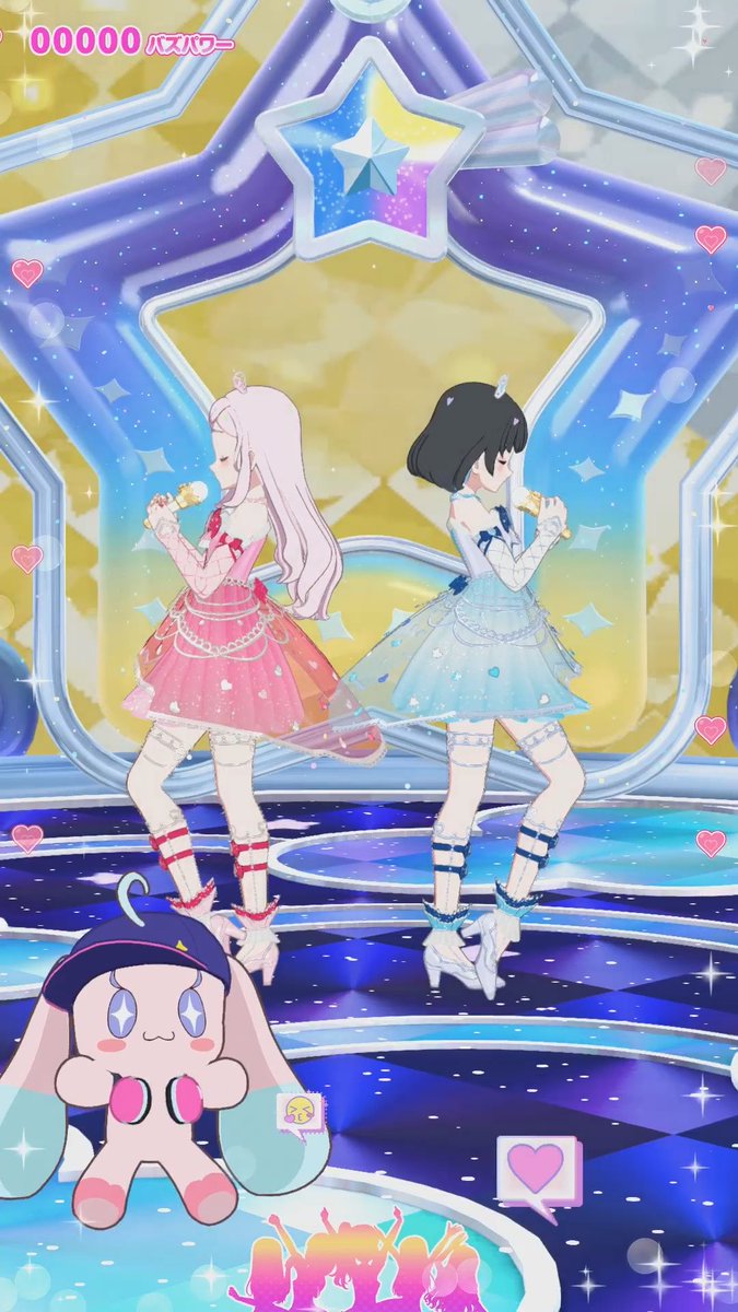 An animated mobile game screenshot showing two anime-style idol characters (one in pink, one in blue) singing on a star-shaped stage with a cute mascot character in the foreground; Japanese text 'パズパワー' appears at the top.