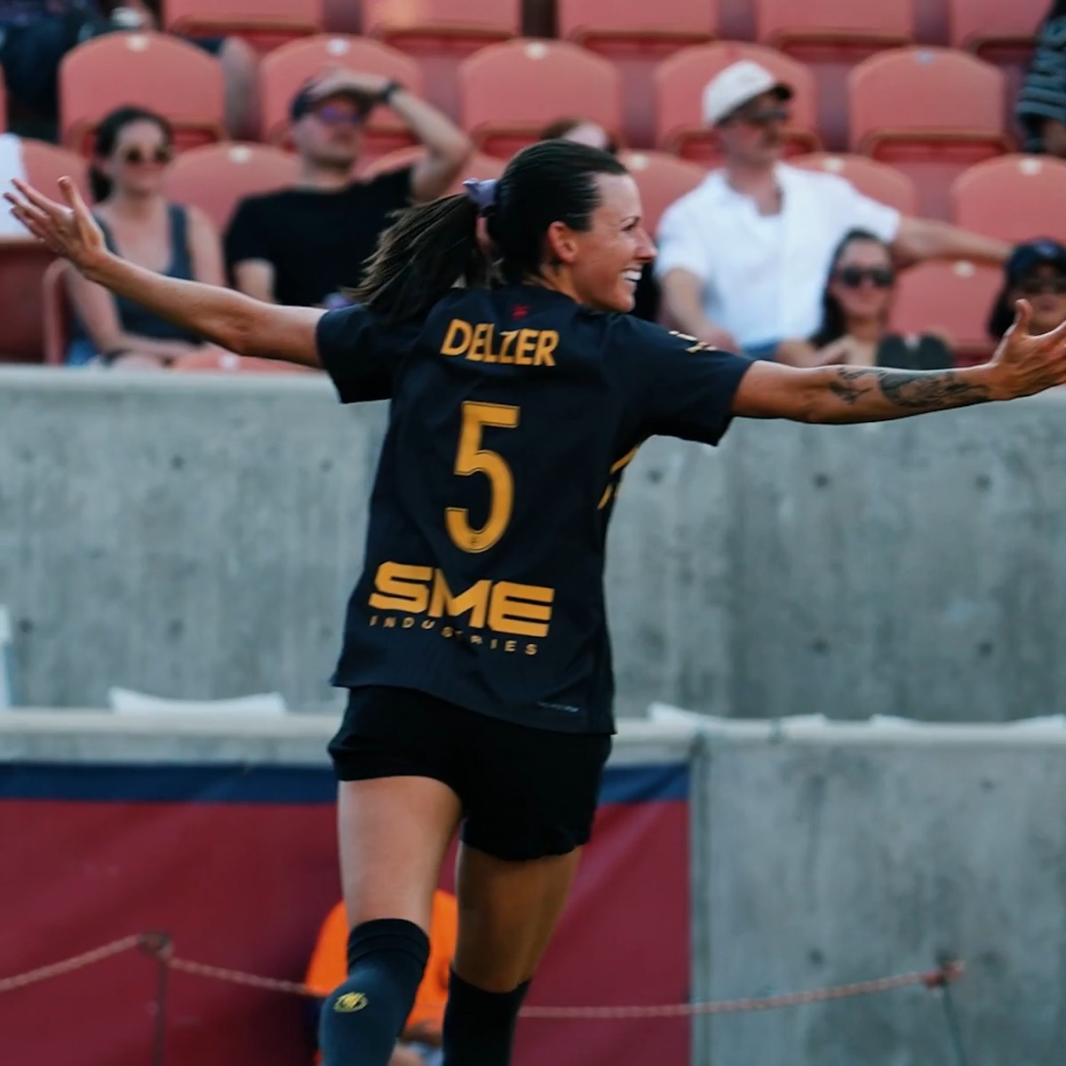 A female soccer player wearing a black jersey with 'DALZER', the number 5, and 'SME INDUSTRIES' printed on the back, celebrating with her arms outstretched in front of stadium seating.