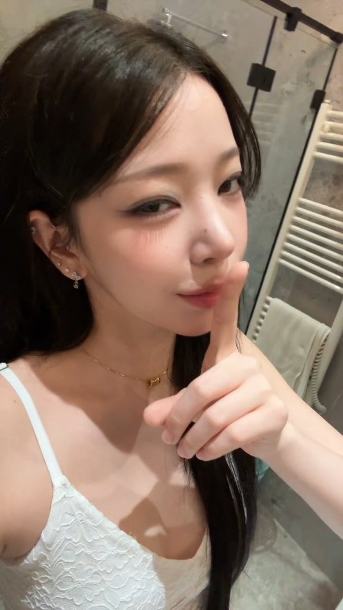 A young woman with long dark hair wearing a white lace camisole and gold necklace takes a close-up selfie in a bathroom.